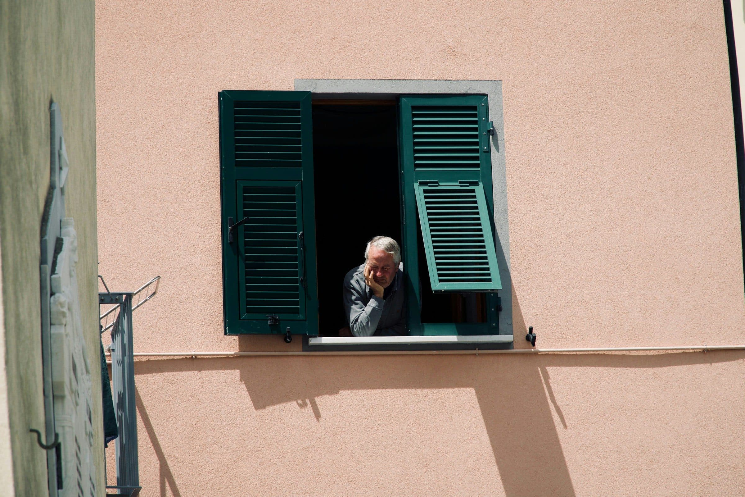 An older man looking out from the shuttered window of a pink house